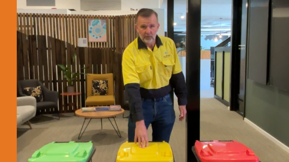 A man in a yellow high-visibility shirt stands in front of three kerbside bins. He is pointing at the bin with the yellow lid.